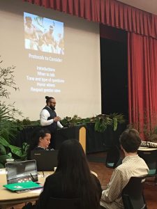 Snapshot from Center for Pacific Island Studies conference. Man with black hair tied in bun wearing white collared shirt and black vest speaking behind podium with powerpoint showing in background. Audience of three is in front. 