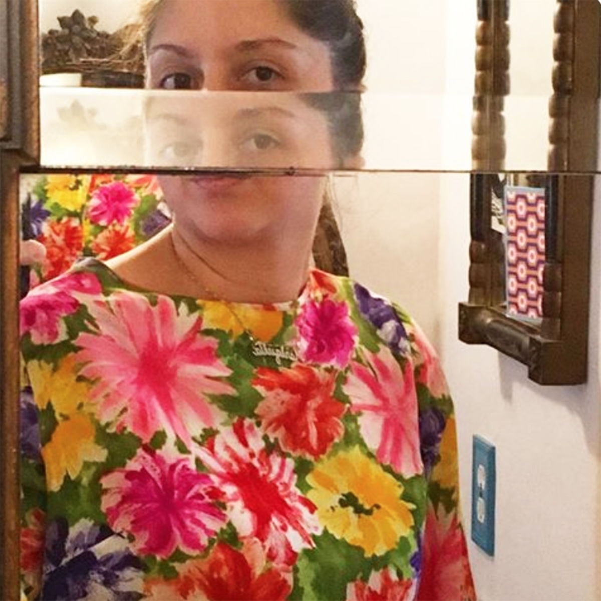 Image shows a photographic portrait of TCP 30-1 featured artist Micki Davis. It is a self-taken photo in front of a mirror, which has distorted Micki's face so that she appears to have two sets of eyes. She wears a flower-print shirt with pink, red, and yellow flowers