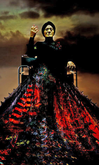 This is a photographic image by TCP 29-1 featured artist Lisa Reihana. It depicts Mehuika, the Maori goddess of fire, as an older woman, sitting on a chair, dressed in a black shirt with a skirt-like item that also appears to be lava