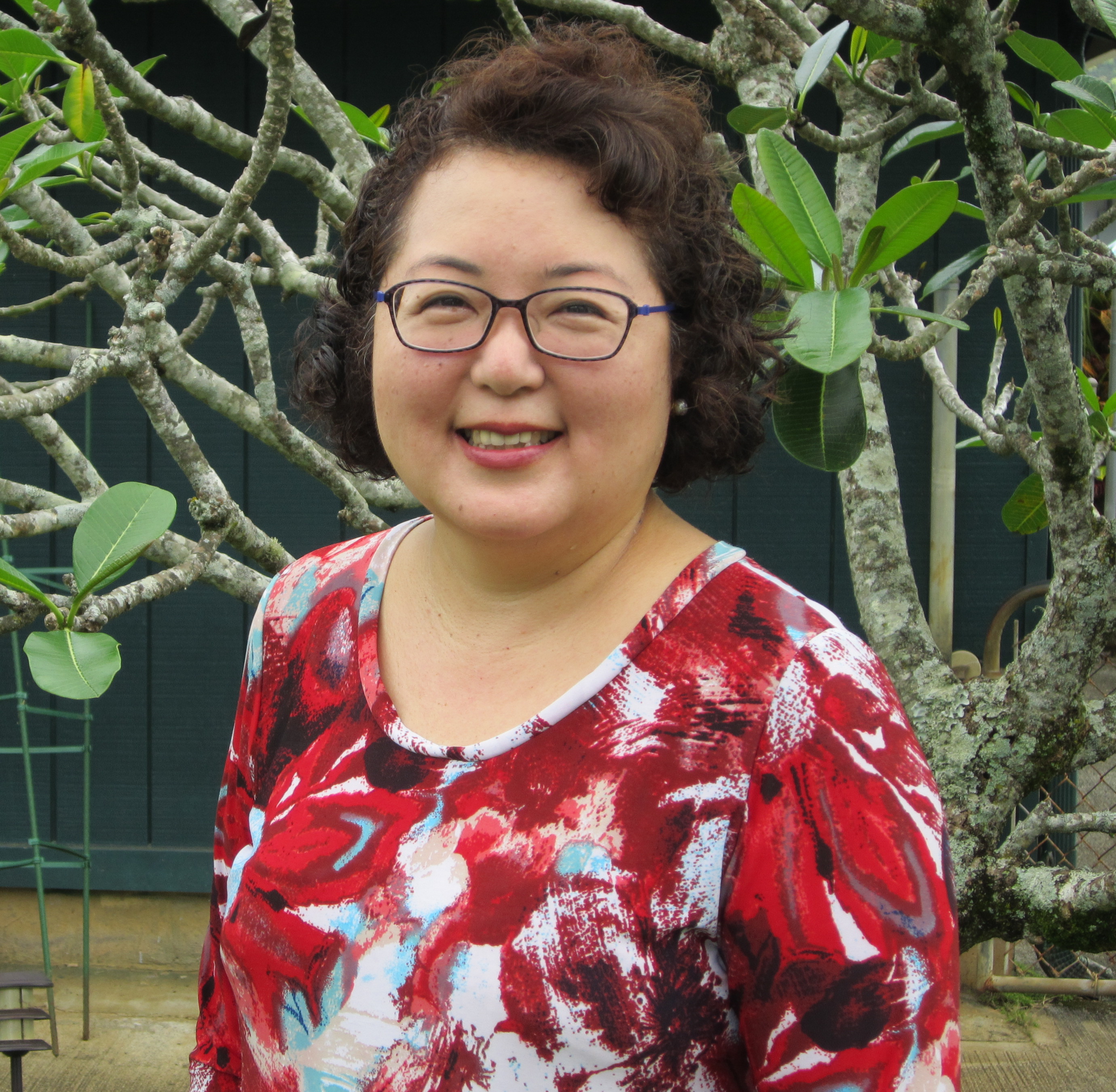 Dr. Satsuma wearing a red, blue, and white blouse and glasses with black and blue frames in front of a trees and a dark green background.