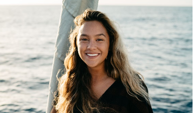 Addie with ocean background. Woman with long wavy hair smiling wearing black v-neck leaning on palm tree.