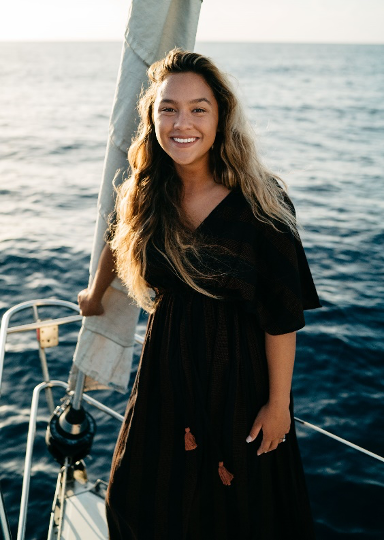 Adeline Ruiz wearing black v-neck dress with long hair smiling. Background has ocean and she is on a boat holding onto a white sail.