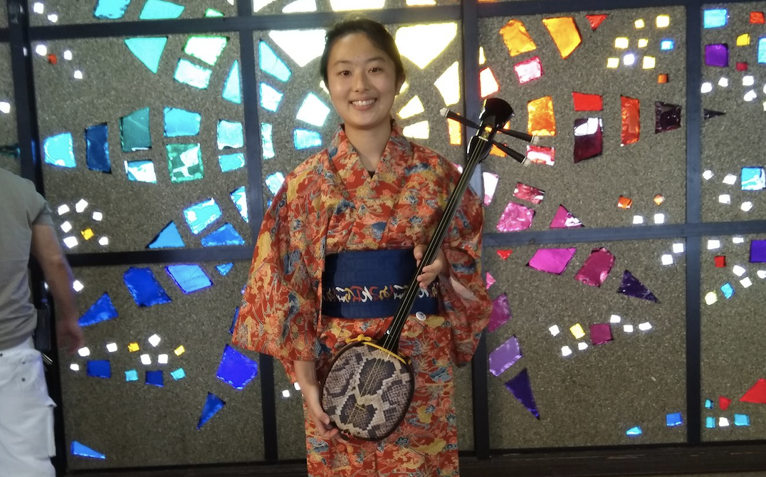 Iroha Mochida smiling and wearing orange-pattern kimono holding Japanese string instrument. Background with colorful tiles in the shape of a sun.