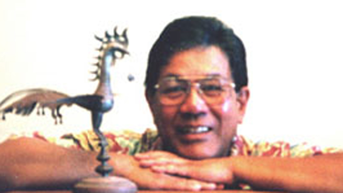 Man with short black hair and glasses wearing a red and yellow collared shirt, Ricardo Trimillos, smiling and placing his heads on top of hands which are on top of one another. Left side has gray bird statue.