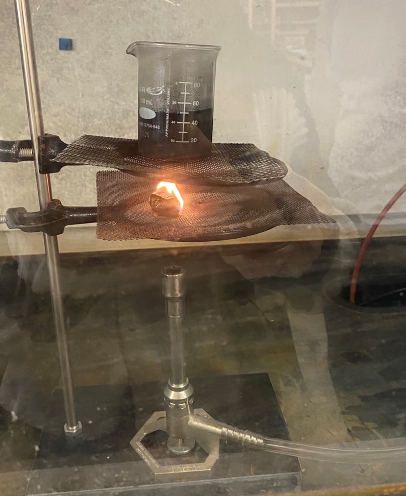 Photograph of a kukui nut on fire from a burner that sits below it. Above the kukui nut is a container with water to measure the heat transfer.