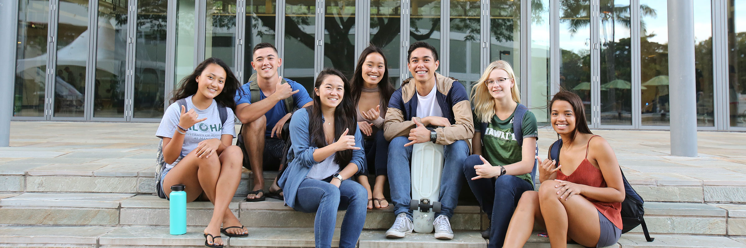 seven students sitting on the ground in front of the WAC Center waving shaka to say hello with smiles on their face