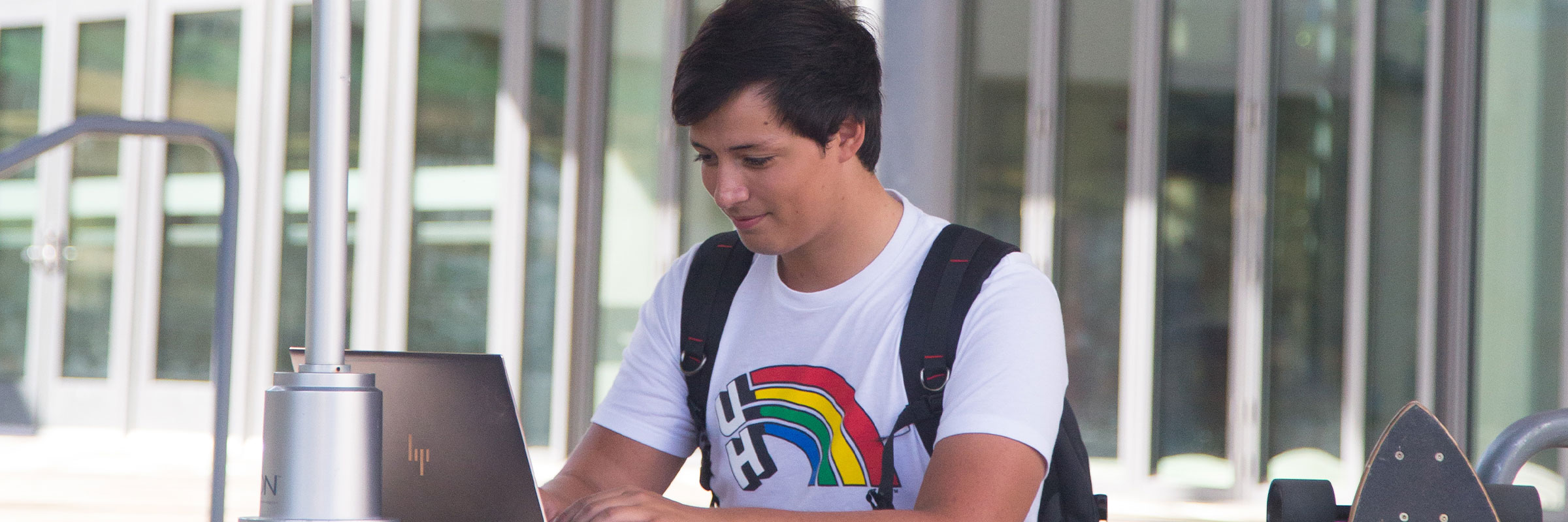 A male student in University of Hawaii T-shirt sitting at a court yard and looking at his computer with a smile on his face