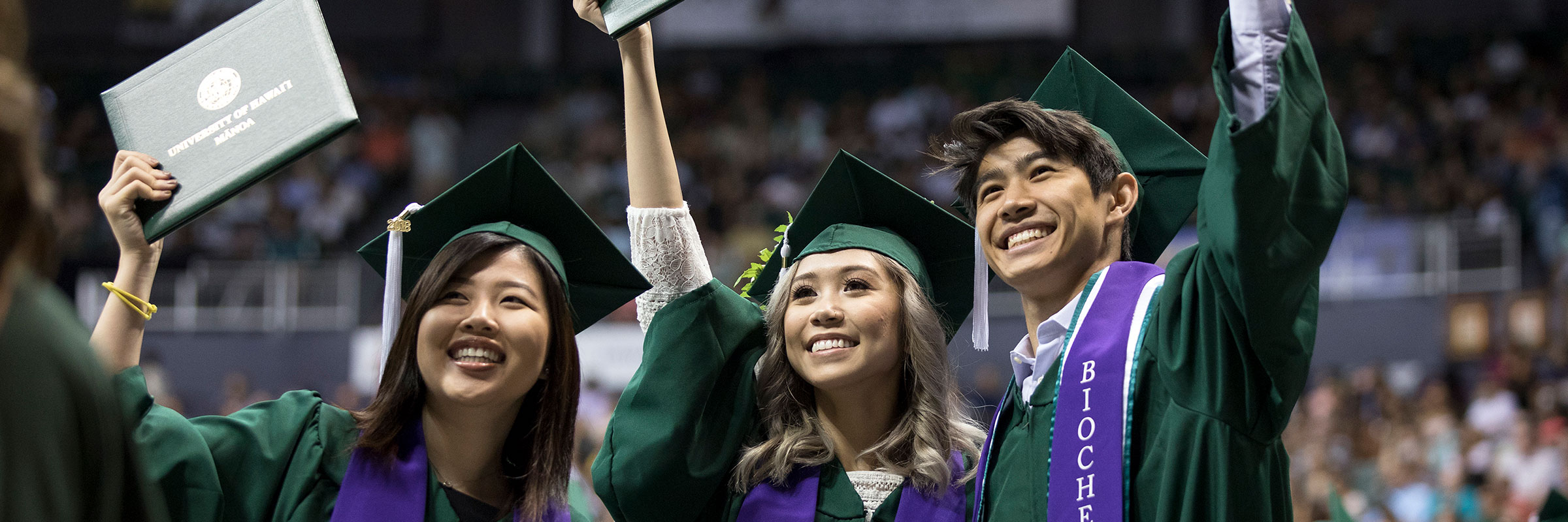 three students in dark green graduation gowns smiling and holding their diplomas at the graduation ceremony
