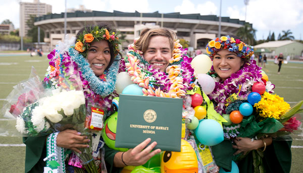 three graduating students dressed in green gown smiling at the camera with flowers lei around their necks