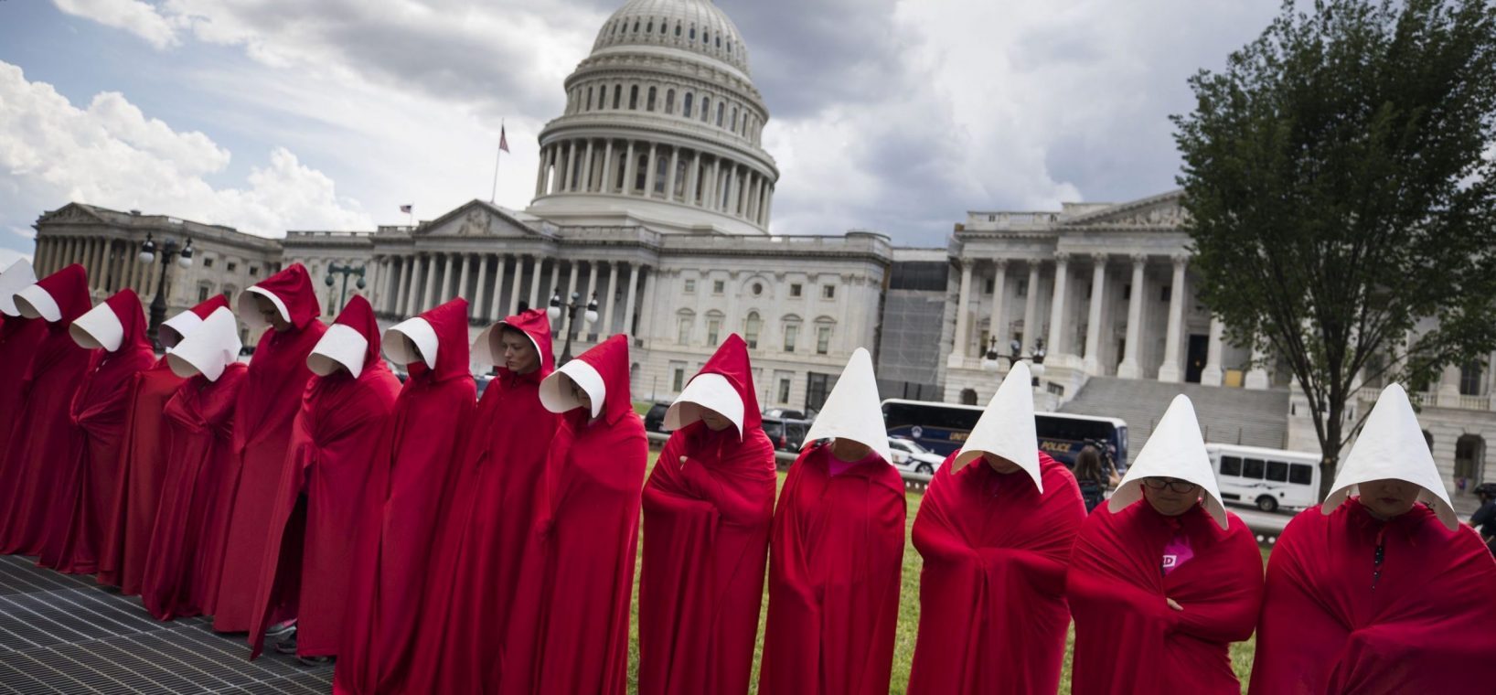 30 women stand somberly in front of the Capitol building in Washington, D.C., dressed in red cloaks and winged bonnets inspired by the Hulu TV adaptation of Margaret Atwood’s novel The Handmaid’s Tale