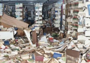 Color image of a library that has been flooded, with books and shelves strewn across the floor.