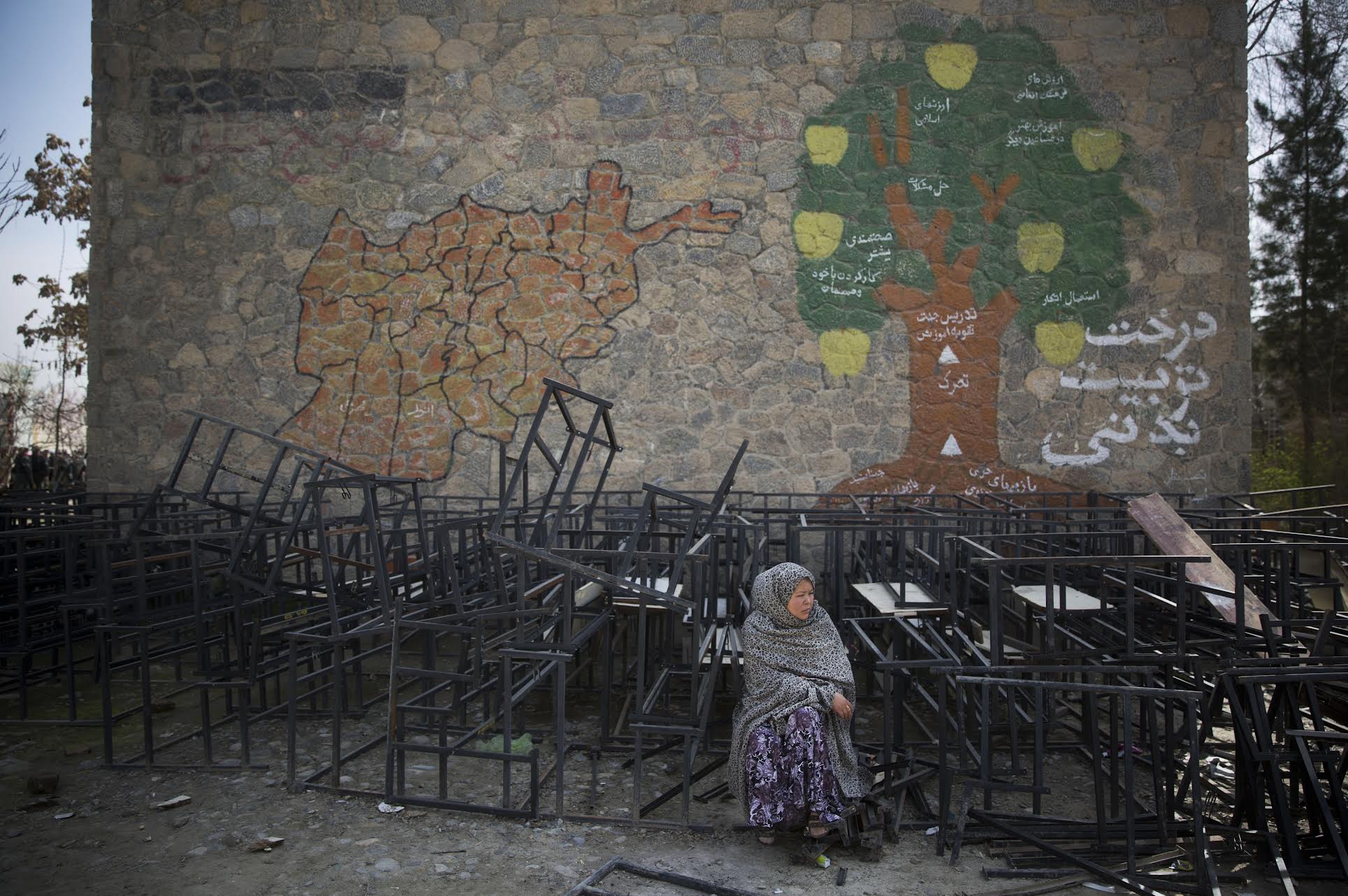 A woman sits among discarded tables and benches. From the Afghanistan: “Graveyard” of Empires Exhibit Flyer
