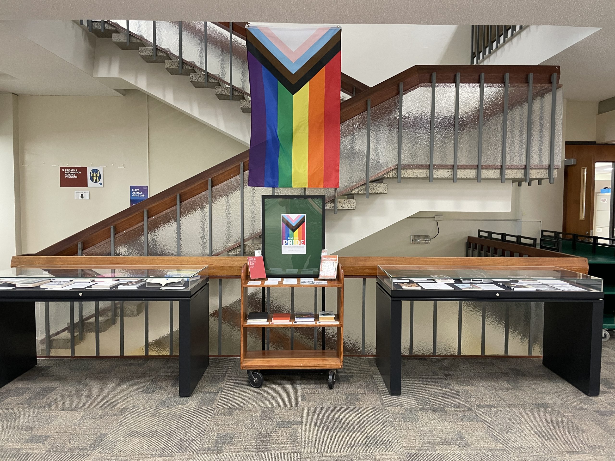 Read with Pride Exhibit, A rainbow flag above 2 display cases and a book cart filled with books