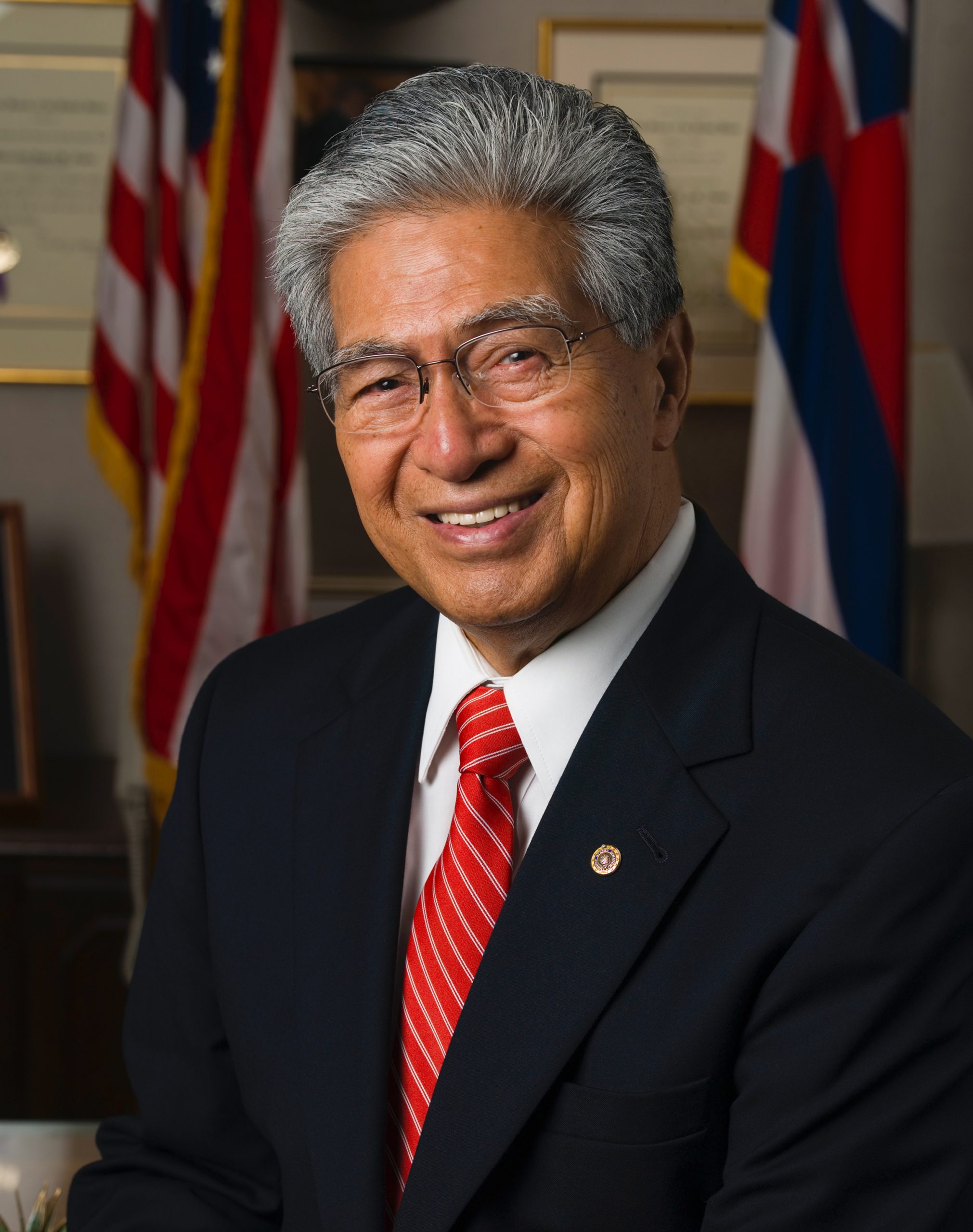 Official portrait of Senator Daniel K. Akaka, in black suit with red tie, in front of U.S. and Hawaiʻi flags.