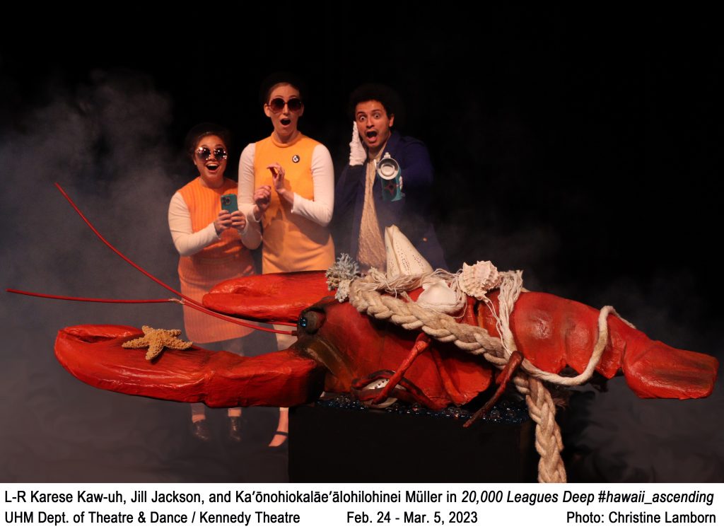 3 college actors holding a giant lobster, 2 wearing matching outfits. Text: L-R Karese Kaw-uh, Jill Jackson, and Ka’ōnohiokalāe’ālohilohinei Müller in 20,000 Leagues Deep #hawaii_ascending. UHM Dept. of Theatre & Dance / Kennedy Theatre           Feb. 24 - Mar. 5, 2023                  Photo: Christine Lamborn.