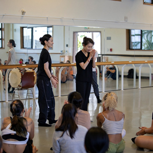 Two Guest Dance Artists From Kora Speaking To Seated Dancers In Front Of Mirror Wall With Barre In Dance Studio.