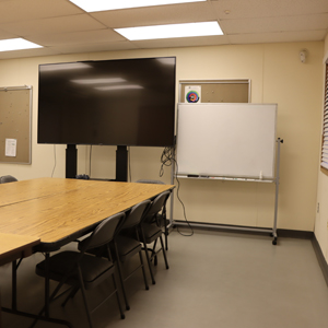 Room With Conference Tables Pushed Together And Surrounded By Chairs. Large Monitor At Foot Of Table. Rolling White Board. Dance Marley Flooring.
