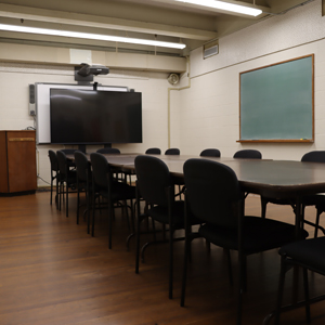 Room With Large Monitor At Foot Of Several Conference Tables Pushed Together And Surrounded By Chairs. Podium. Chalkboard. Hardwood Dance Floor.