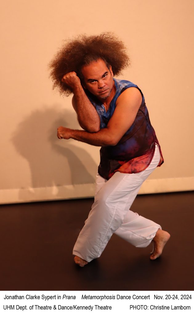 Male dancer with afro pulled back in blue and red top with white pants in a strong hip-hop pose looking at camera.