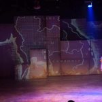 A female performer in a head scarf stands to the right, in front of a projection of a map of the region of Southeast Asia centered around Thailand, which labeled here as Siam