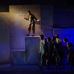 A group of performers moving across the stage in dim blue and yellow lighting, with one actor above the rest standing on a tall block