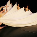 Dancer wrapped in white fabric with edges pulled out by four other dancers