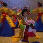 Dancers in bright costuming; center dancer squatting holding flowers; more dancers circle around