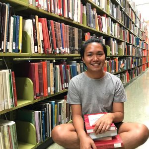 UROP student, Jeremiah Bonilla, in a library with a stack of books needed for her project.