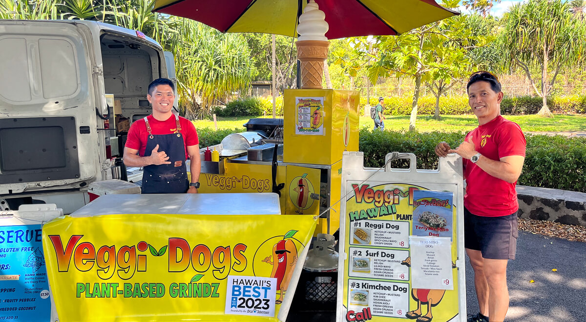 Two smiling employees standing beside the Veggie Dogs mobile food truck, serving plant-based cuisine