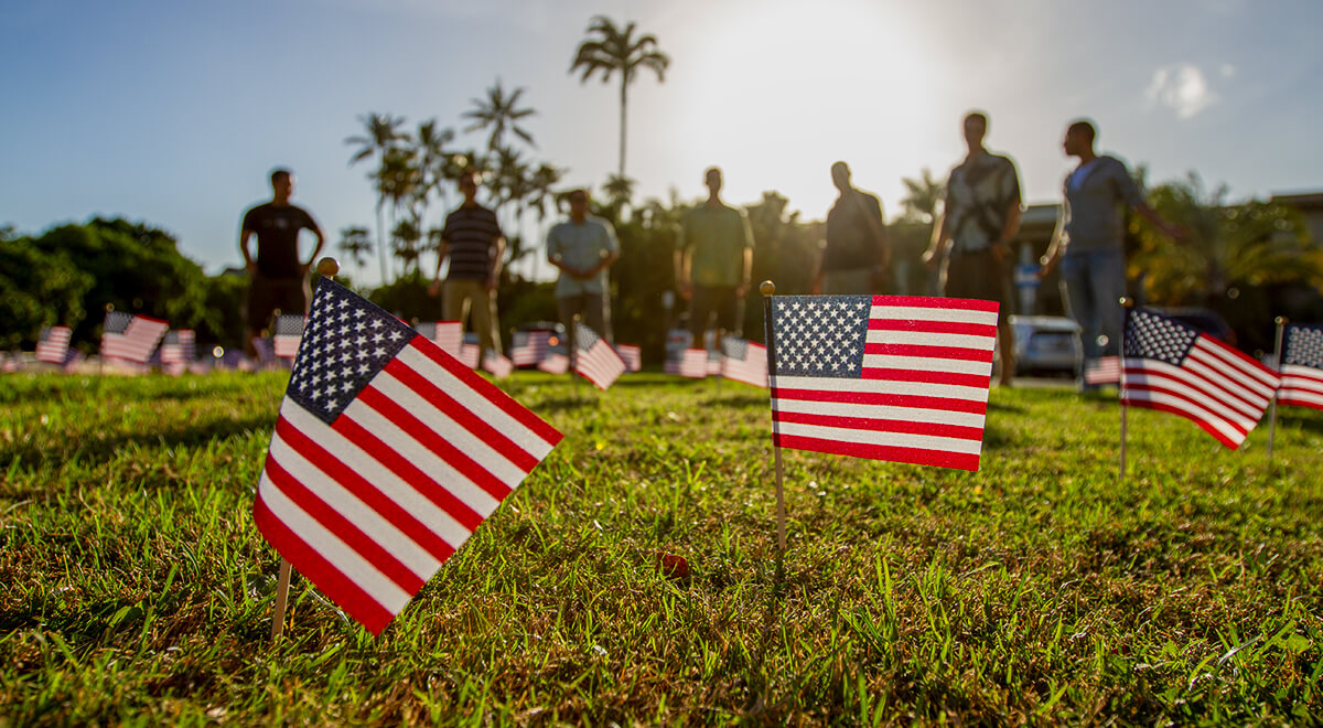 Small American flags posted in the Varney Circle lawn, strongly backlit by the late afteroon sun, and seven out-of-focus veterans standing in the background