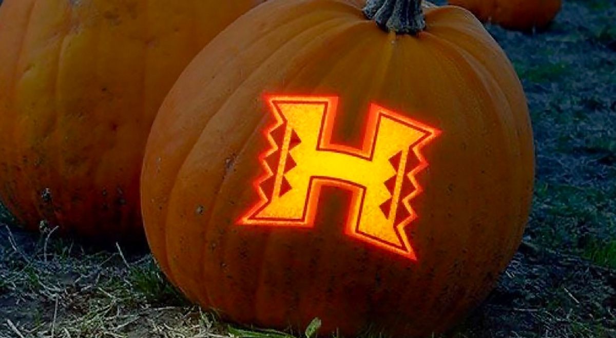 Two pumpkins on the ground at dusk with the glowing Hawaii Warriors H logo cut into one